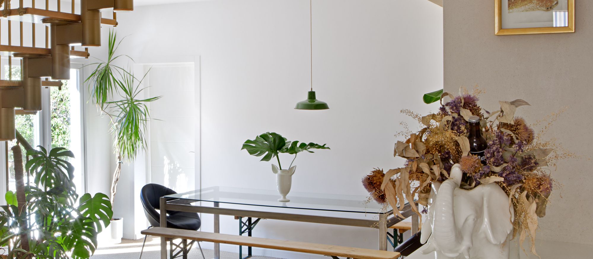 Comedor luminoso con mesa de cristal, banco de madera y plantas junto a un gran ventanal